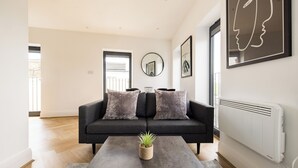 A modern living room with a grey sofa, two matching cushions, a coffee table with a plant, a round mirror, and framed artwork on the wall. - Staines Riverside by Charles Hope (Staines)