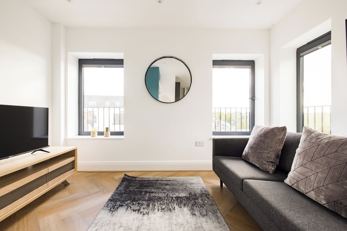A modern living room with a flat-screen TV, a grey sofa with patterned cushions, a round mirror, and two windows with a view of the outside.