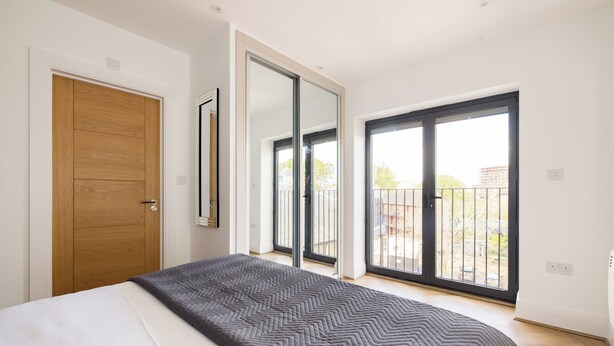 A bedroom with a large bed, a wooden door, a sliding glass door to a balcony, and a view of the outside. - Staines Riverside by Charles Hope (Staines)
