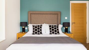 A neatly made bed with a grey blanket, two black and white patterned pillows, and a brown headboard. There are two bedside tables with lamps and a wooden door in the background.