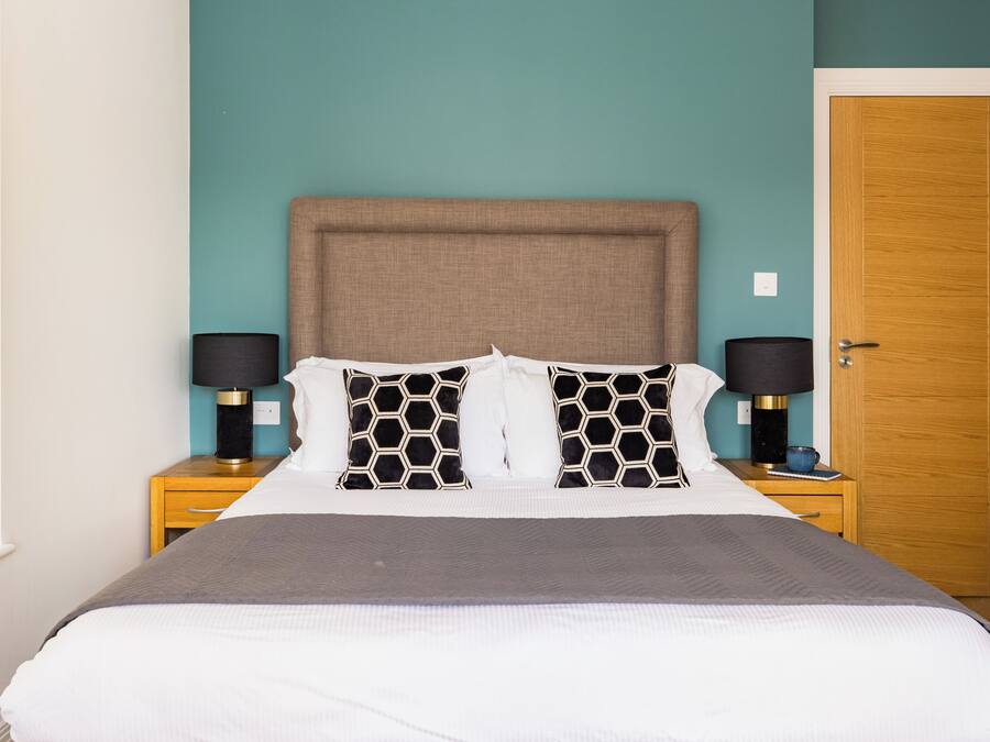 A neatly made bed with a grey blanket, two black and white patterned pillows, and a brown headboard. There are two bedside tables with lamps and a wooden door in the background.