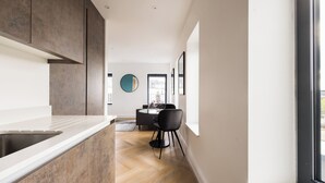 A modern kitchen with a large island, a dining area with a round table and chairs, and a view through a window. - Staines Riverside by Charles Hope (Staines)