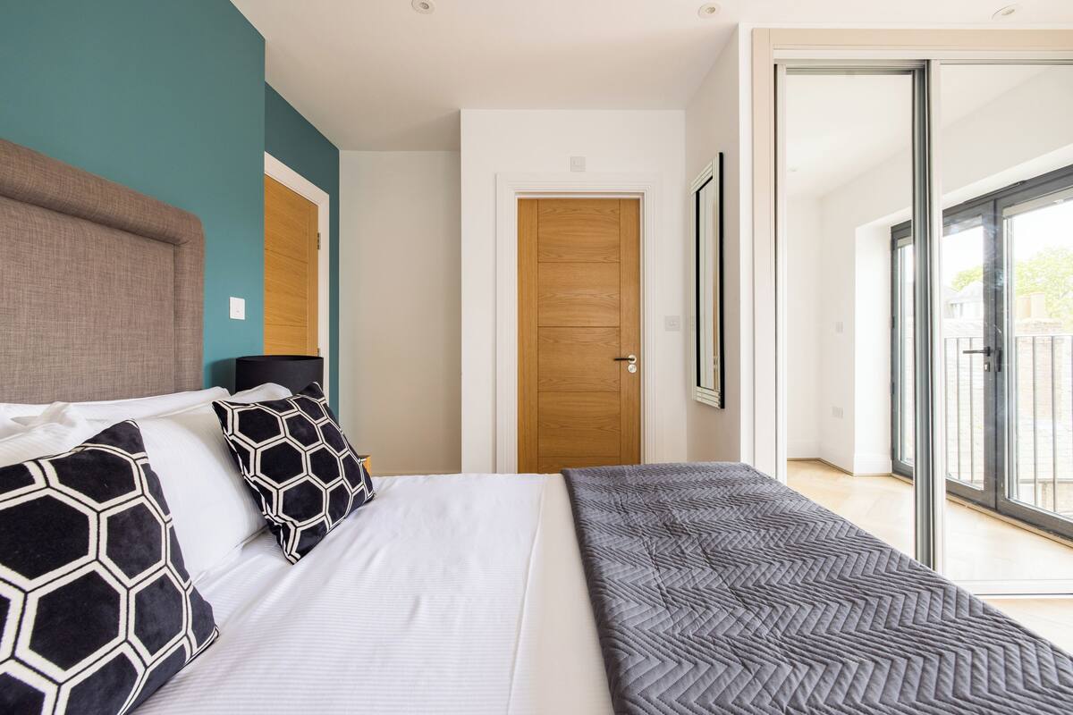 A modern bedroom with a large bed, a wooden door, and a view of the outdoors through a glass door.