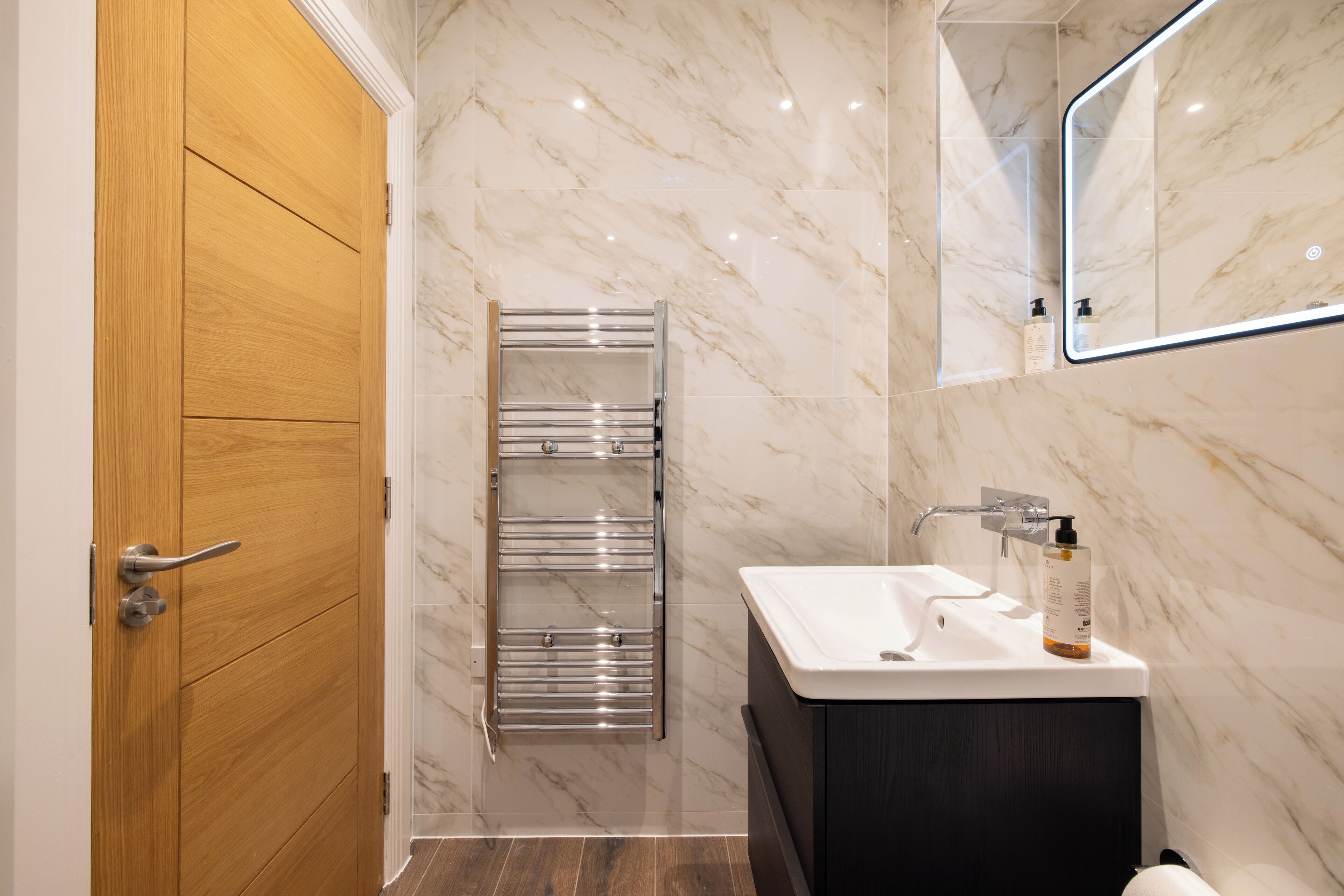 A modern bathroom with a wooden door, a chrome towel rack, a white sink with a black cabinet, and a marble wall.