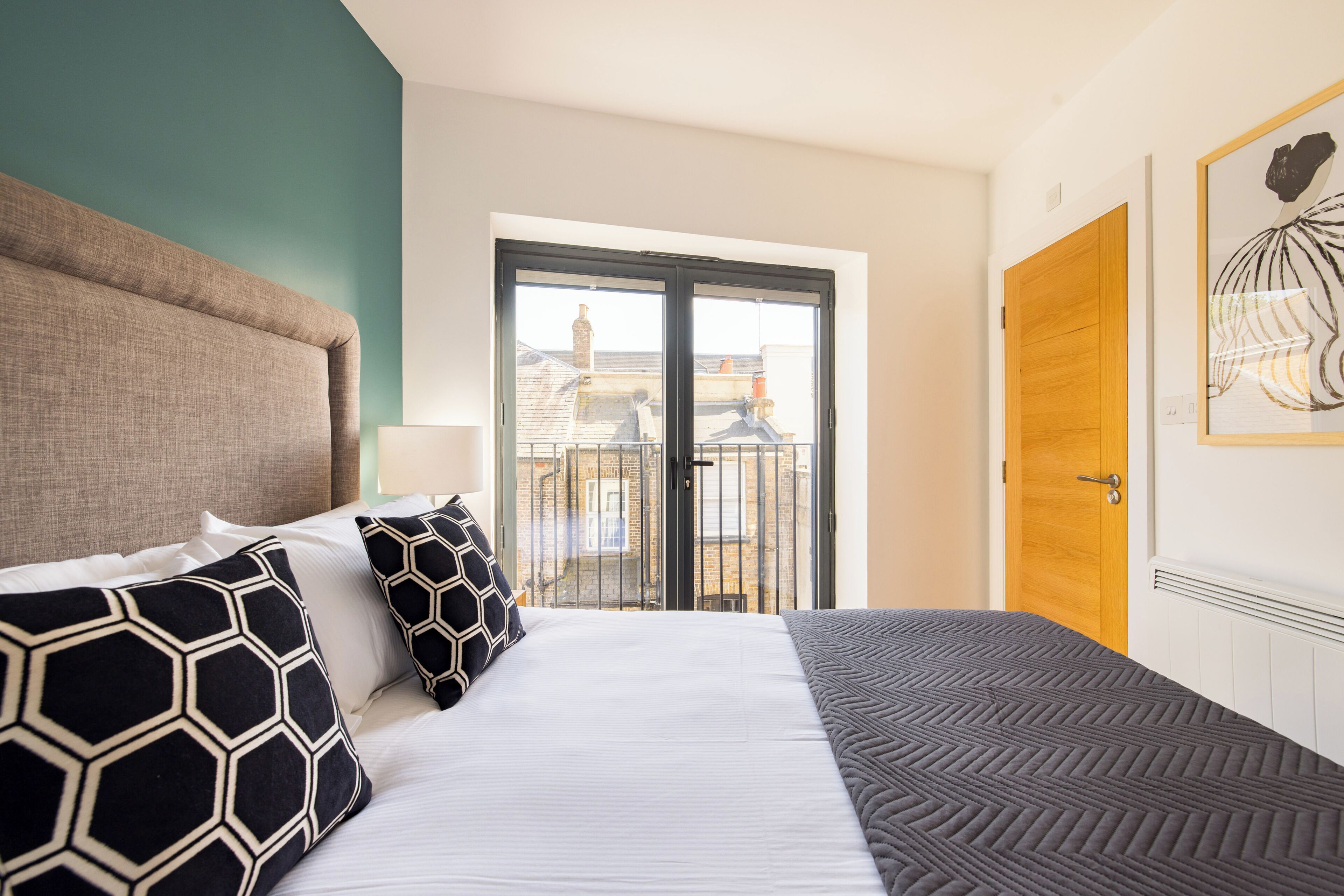 A hotel room with a large bed, a view of the city through a balcony door, and a wooden door.