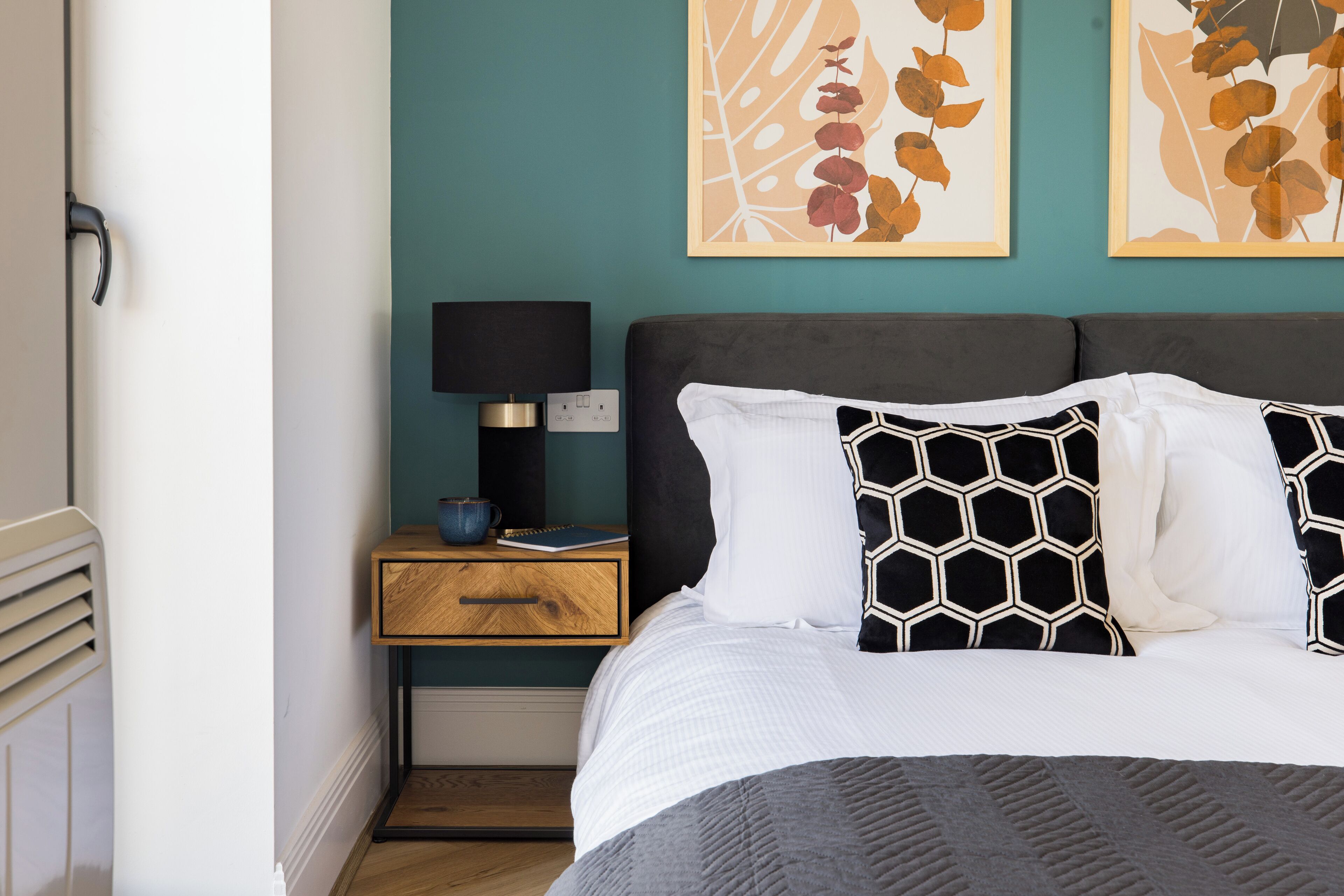 A neatly made bed with a patterned pillow, a bedside table with a lamp, and two framed artworks on the wall.