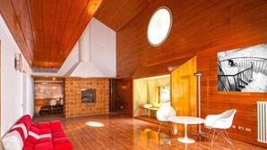 Une pièce spacieuse avec un ensemble de canapés rouges, une petite table et des chaises. On y trouve une cheminée, une grande fenêtre et un tableau accroché au mur.
