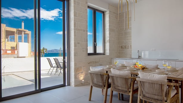 A dining area with a table set for four, a view of the sea through large windows, and a balcony with chairs. - Perla Dream Luxury Villa (Chania)