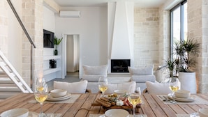 A dining area with a wooden table set for two, white plates, and glasses of white wine. There is a sofa and a TV in the background. - Perla Dream Luxury Villa (Chania)