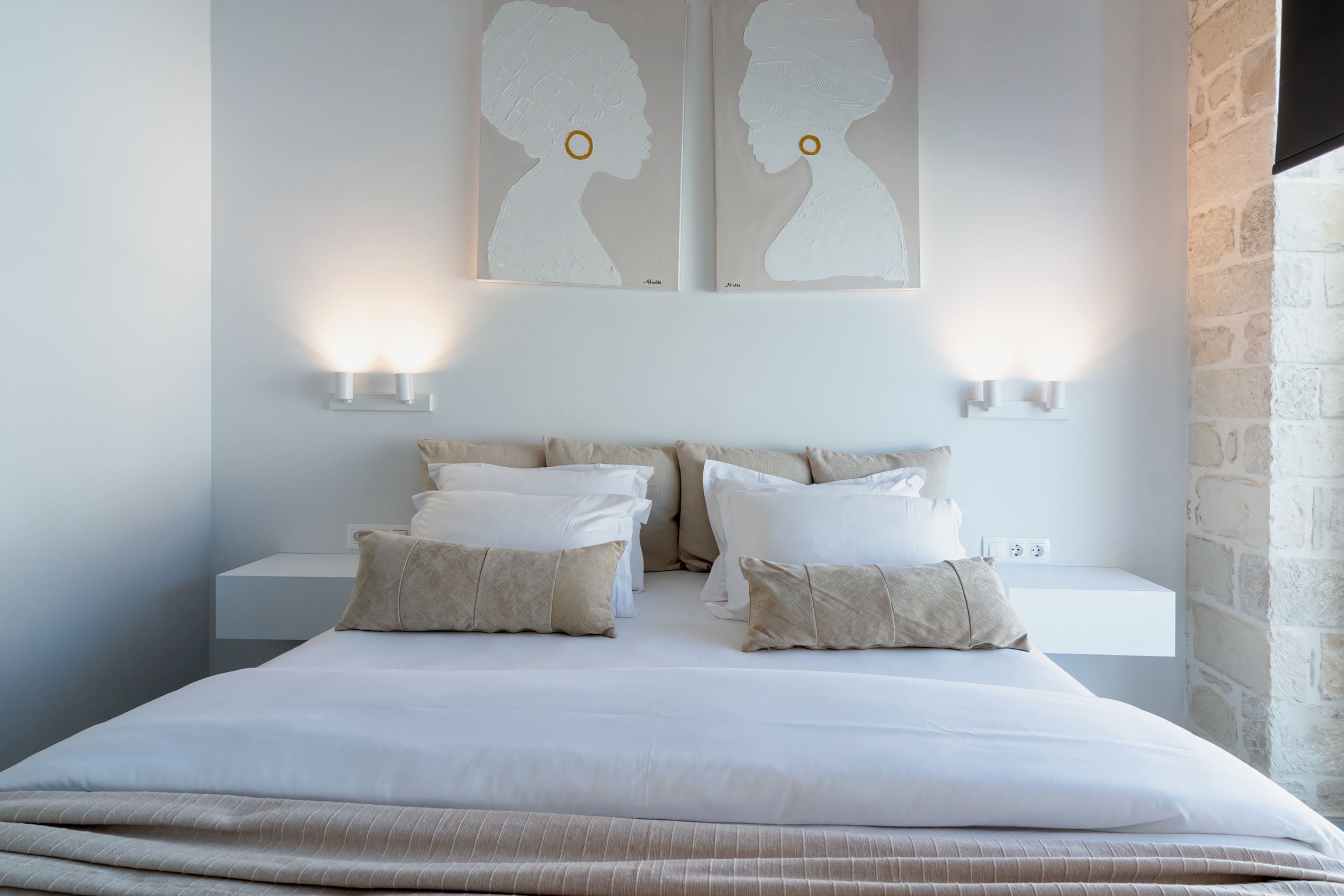 A neatly made bed with white linens and beige pillows, two wall-mounted light fixtures, and a white nightstand.