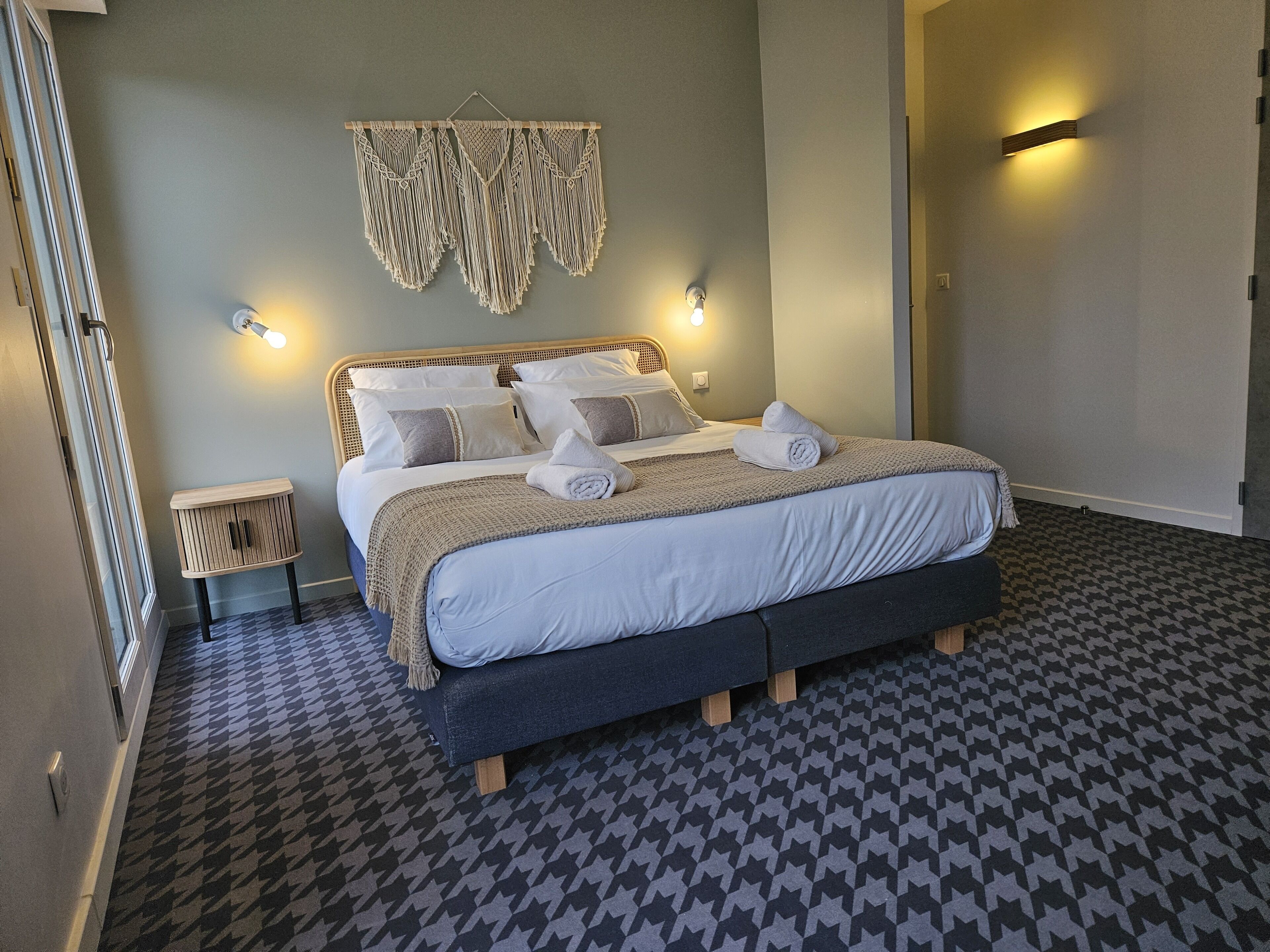 A neatly made bed with a patterned floor, a bedside table, and a macrame wall hanging.