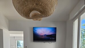Un comedor con una larga mesa de madera dispuesta para una comida, un televisor montado en la pared y ventanas amplias que permiten la entrada de luz natural.