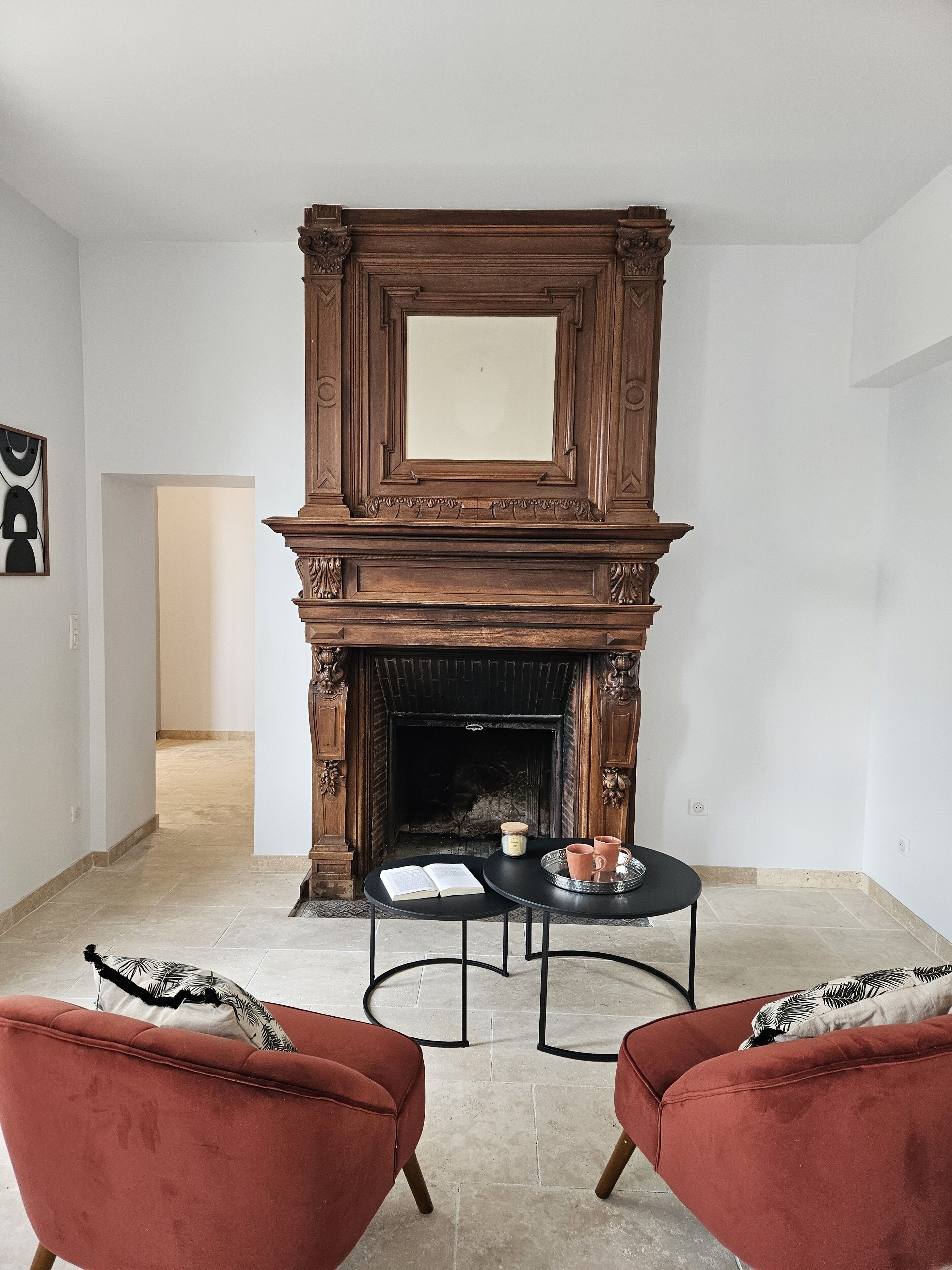 Une pièce avec une cheminée, deux fauteuils rouges, une petite table avec un livre et une bougie, et un tableau encadré au mur.
