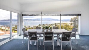 A modern dining area with a large table and chairs, large windows offering a view of a body of water and mountains.