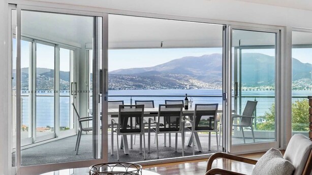A modern living room with a glass door leading to a balcony with a table and chairs, overlooking a scenic view of a lake and mountains.