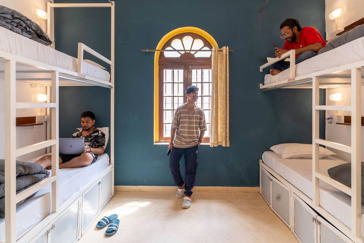 Habitación tipo dormitorio con literas, una ventana de marco amarillo y una persona de pie en el centro.