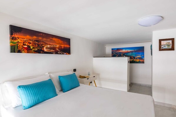A hotel room with a bed, two blue pillows, a nightstand, a water bottle, and two paintings of coastal scenes.