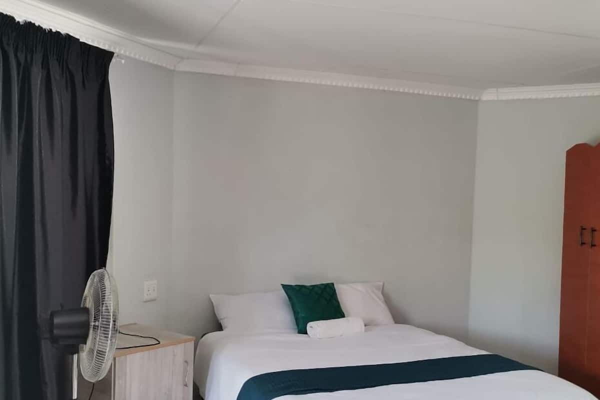 A neatly made bed with white and green pillows, a bedside table with a fan, and a dark-colored wardrobe.