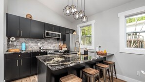 A modern kitchen with dark cabinetry, a central island with stools, a stainless steel microwave, and a window with a view of greenery. - Romantic Beehive Treehouse Getaway in Nature (Pittsboro)