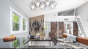 A modern kitchen with a large island, a dining area with a sofa, a staircase, and a view of greenery outside. - Romantic Beehive Treehouse Getaway in Nature (Pittsboro)