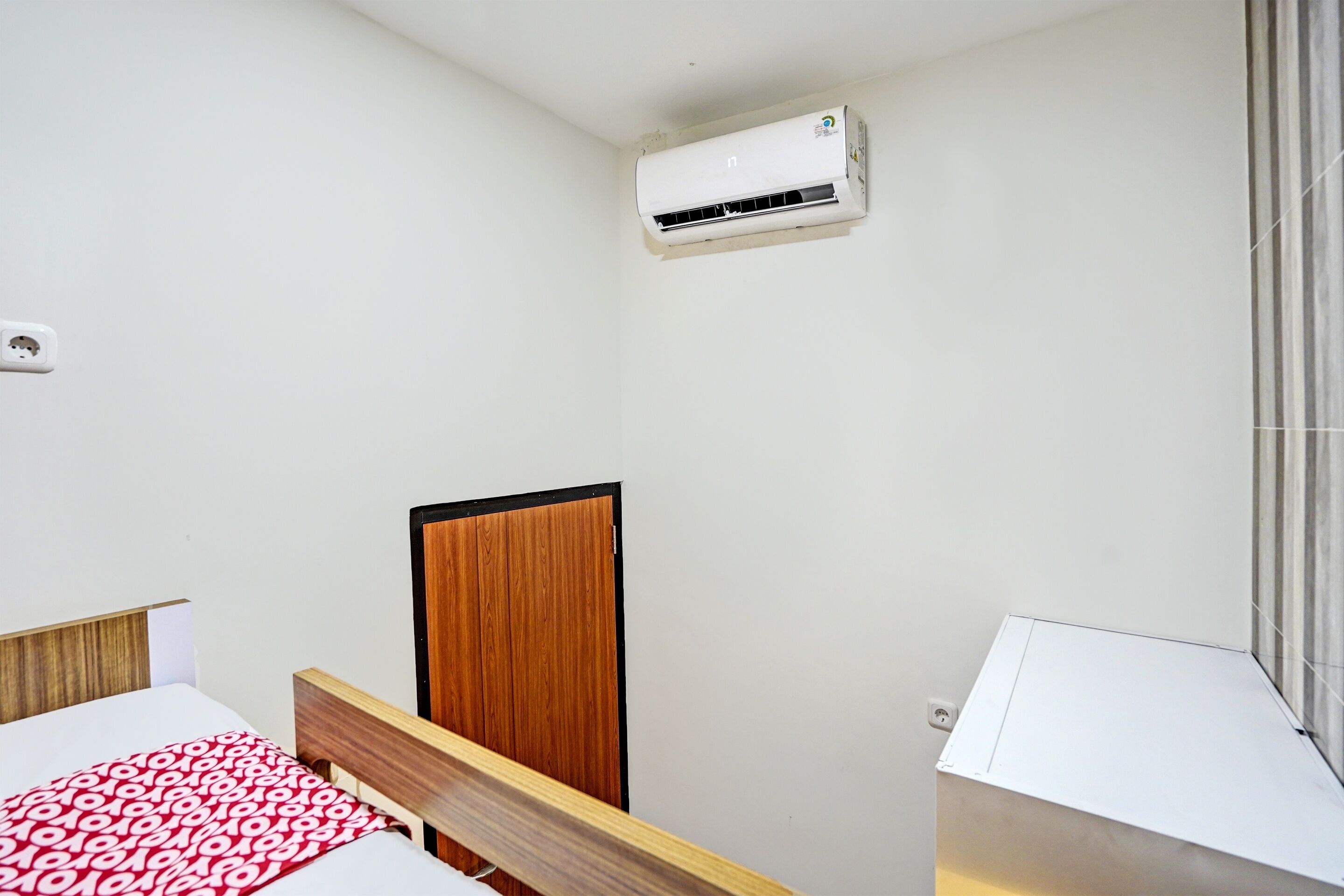 A hotel room with a bed, a wooden wardrobe, an air conditioning unit, and a window with curtains.