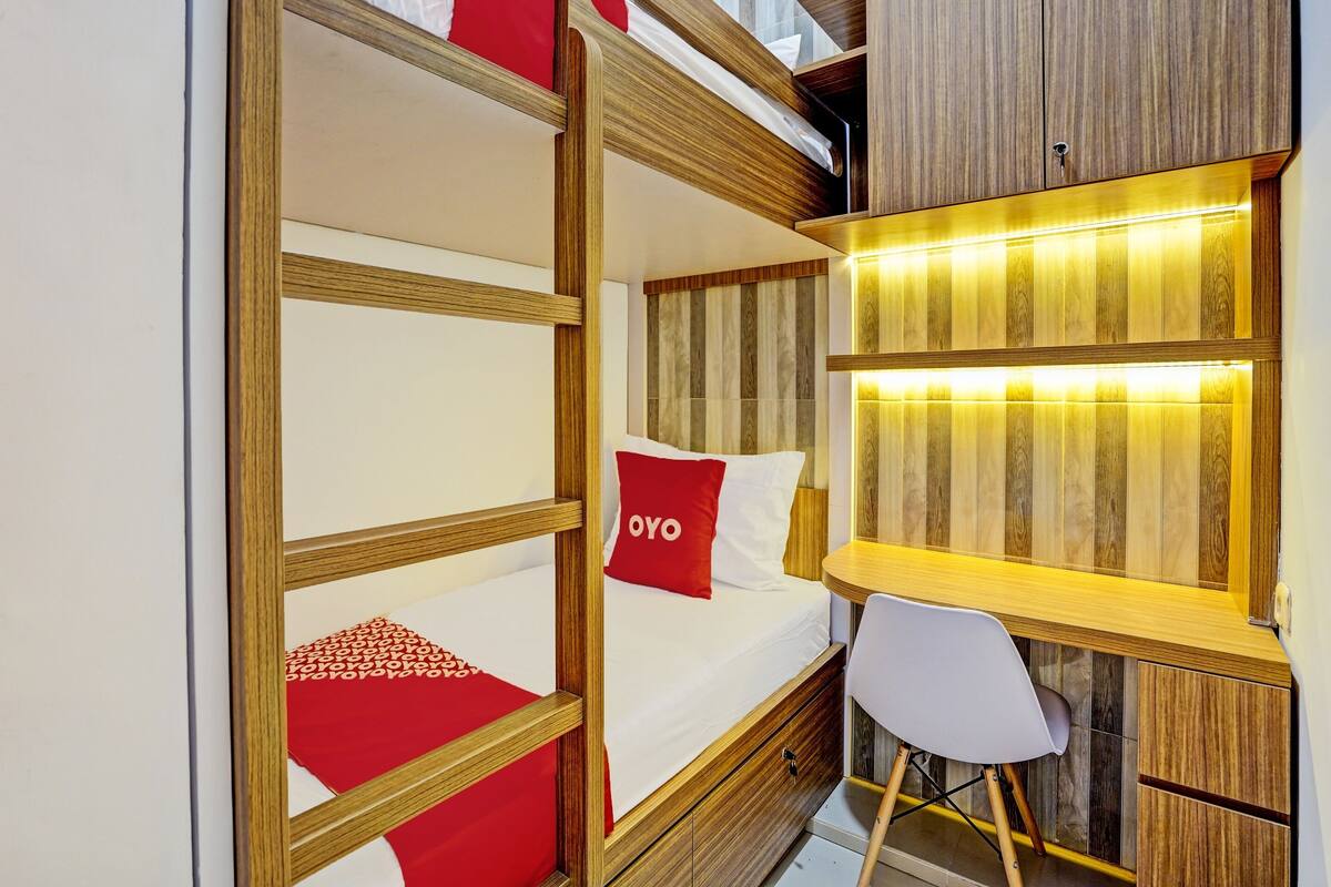 Ein Stockbett mit rotem Kopfkissen, ein Schreibtisch mit Stuhl und ein Holzkasten.