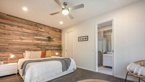 Ein Schlafzimmer mit Bett, Deckenventilator, Nachttisch, Nachttischlampe, einer Wand als Holzdekor, einem Durchgang zu einem Badezimmer und einem Sessel mit Kissen.