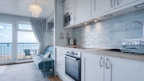 A modern kitchen with white cabinets, a built-in oven, and a microwave. There is a view of the ocean through large windows. - 17 Beach Court - 2 Bed Apartment - Saundersfoot (Saundersfoot)