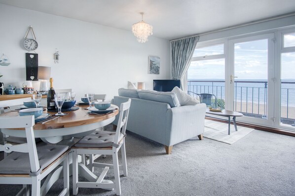 Una sala de estar moderna con comedor, un sofá azul claro, una araña y vista al mar a través de amplios ventanales.