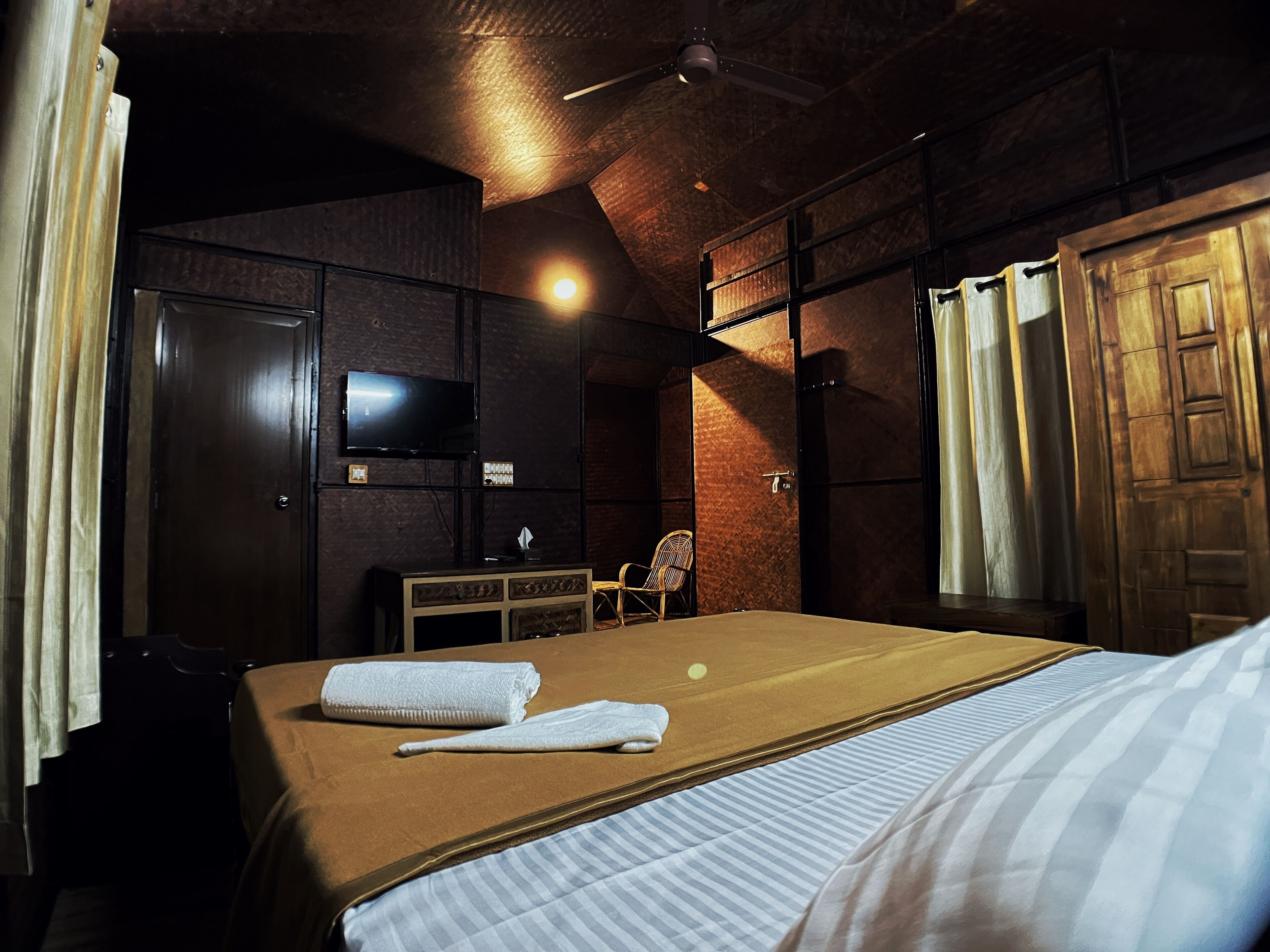 A hotel room with a large bed, a television on the wall, a wooden wardrobe, and a window with curtains.