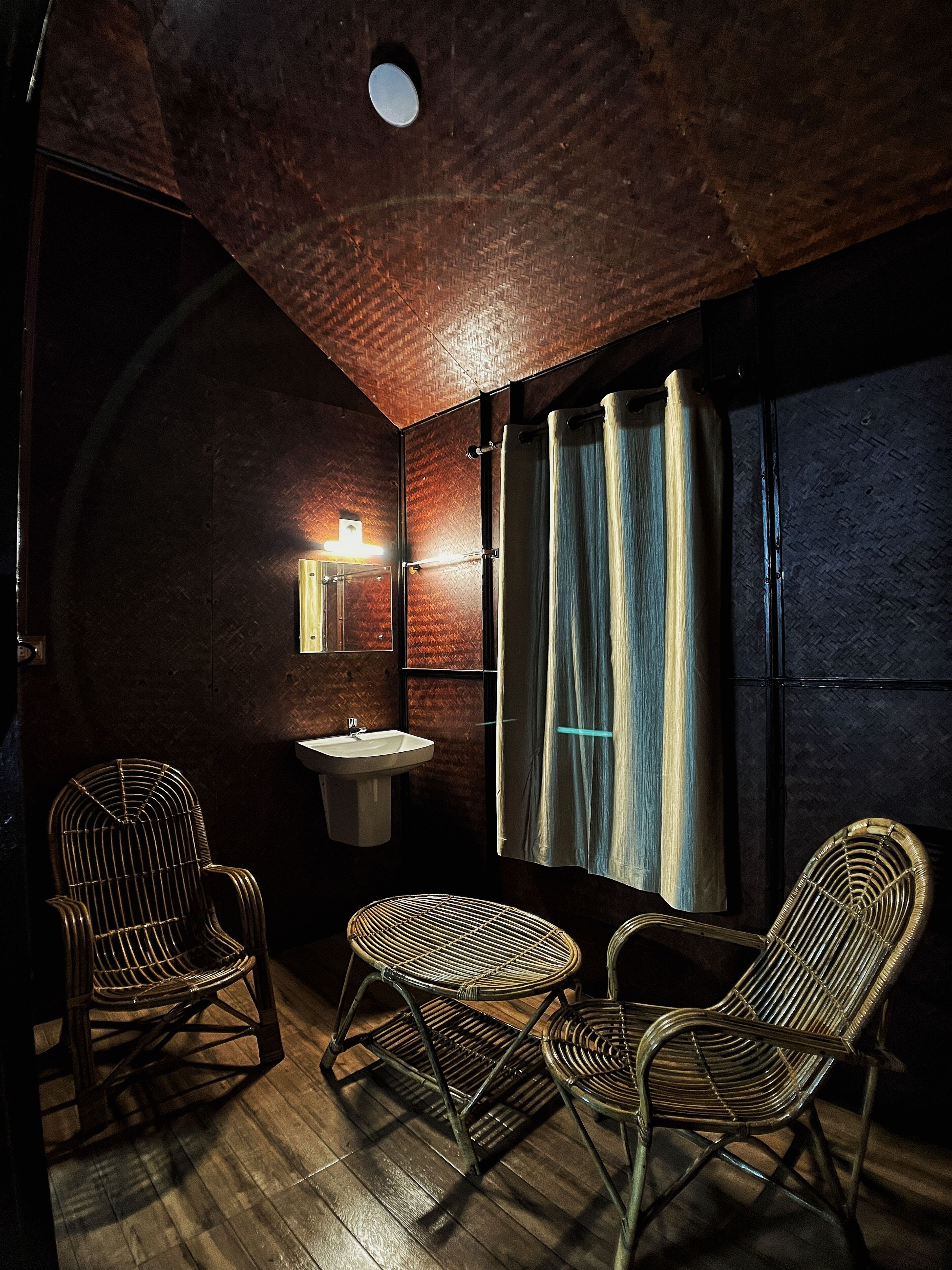 A small, dimly lit room with a wooden ceiling, a single wall-mounted sink, and three wicker chairs.