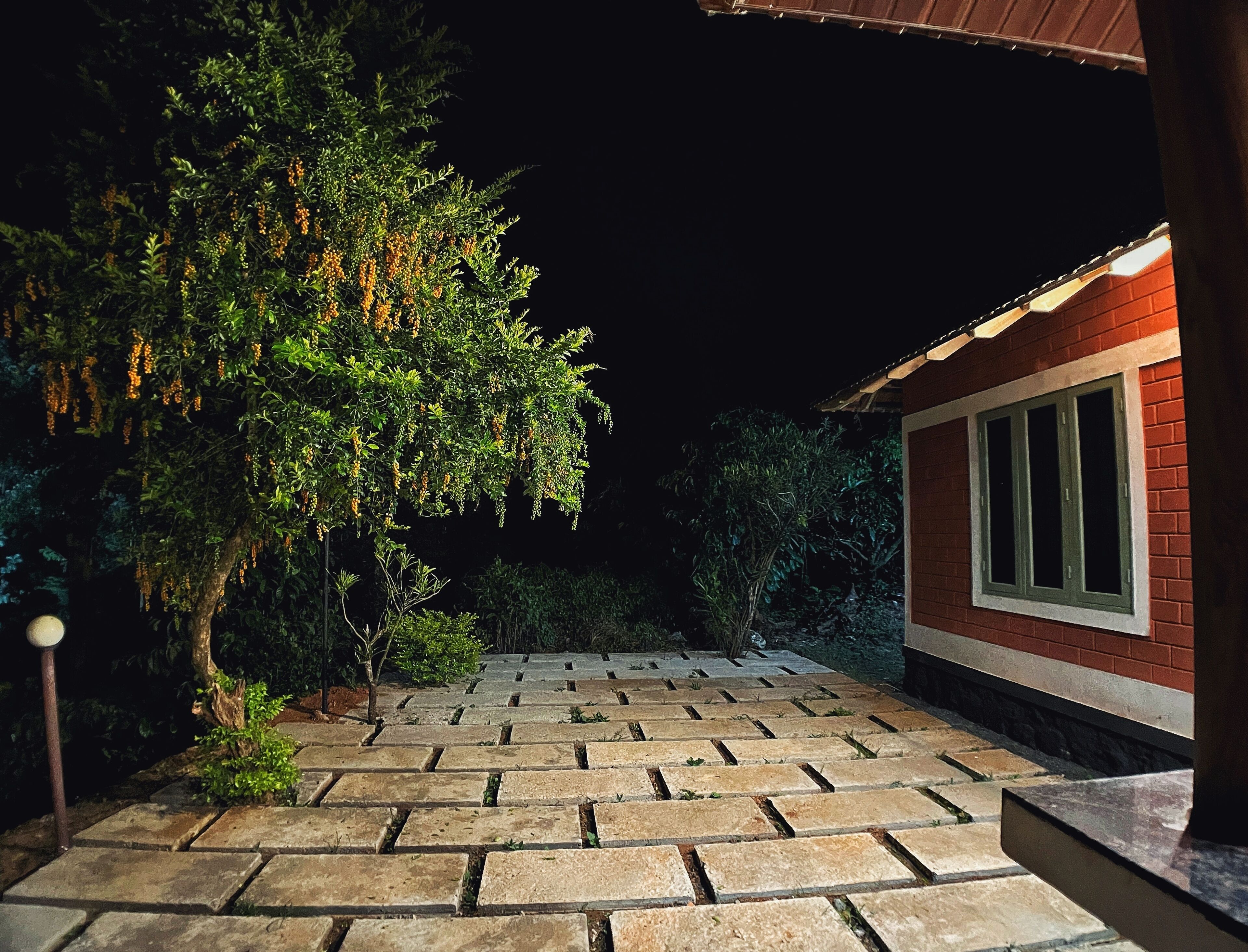 A paved outdoor area with a stone pathway leading to a building with a red brick exterior and a window with green shutters.
