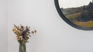A room with a round mirror reflecting a landscape, a small table with a vase of dried flowers, and a teal bench.