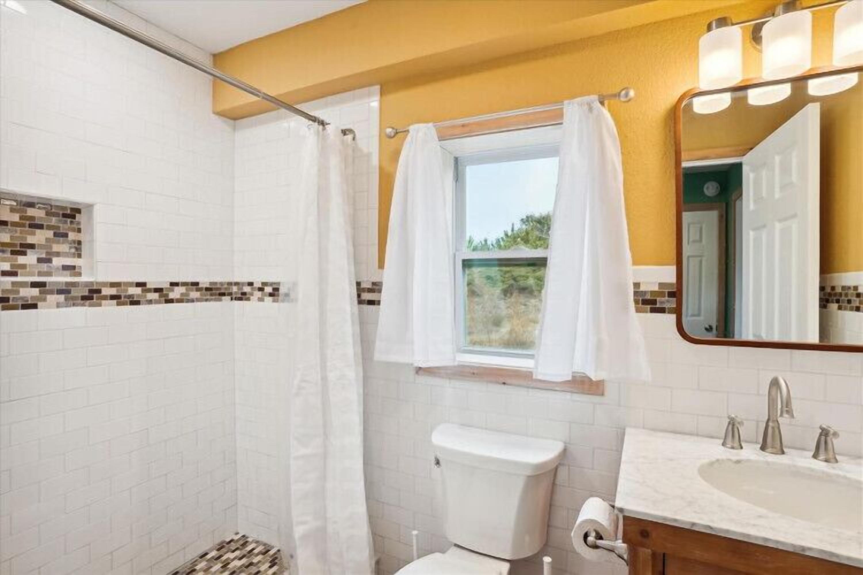 A bathroom with a shower, toilet, sink, and a window with a curtain.