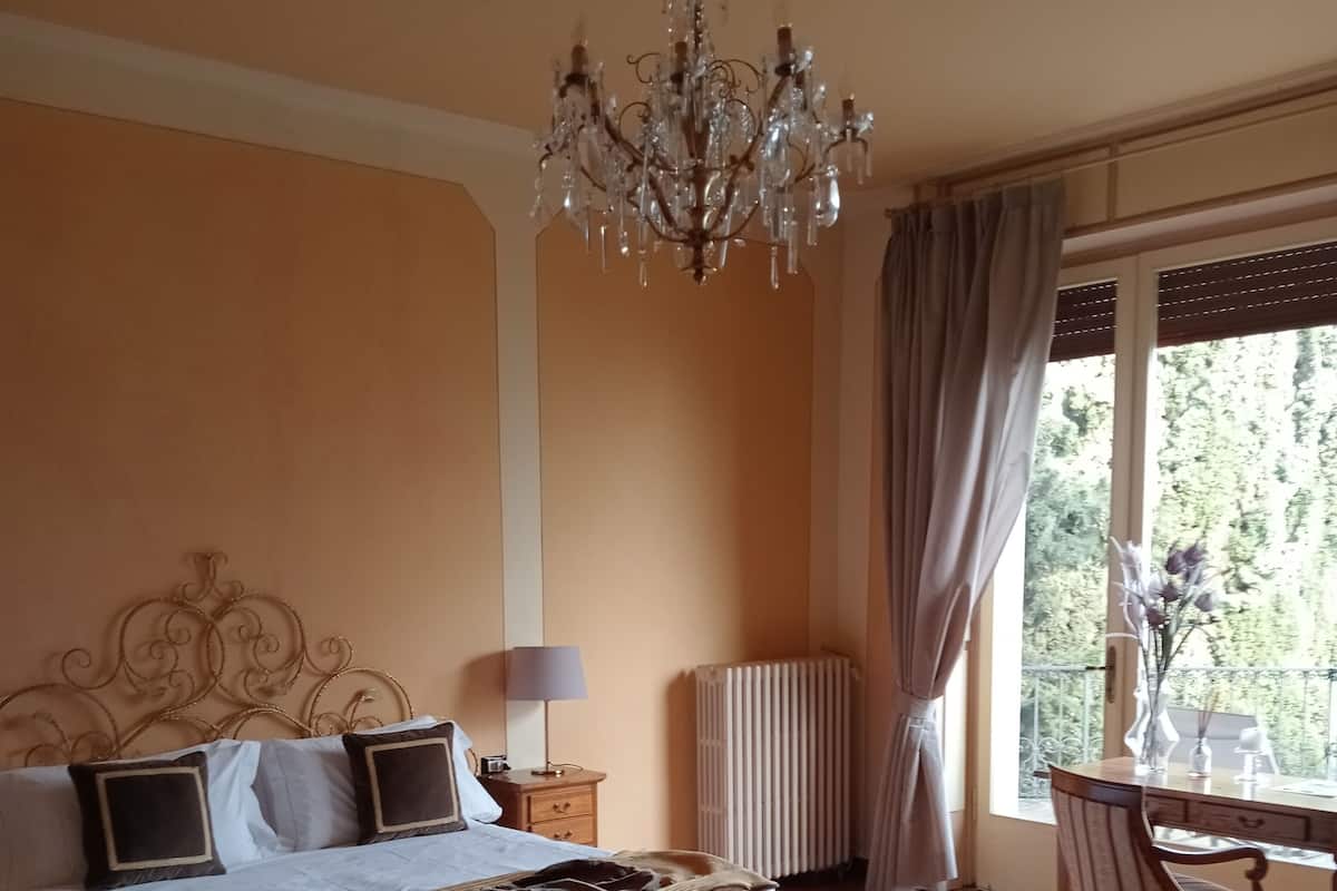 A bedroom with a large bed, a chandelier, a radiator, a dining table with chairs, and a view of greenery outside.