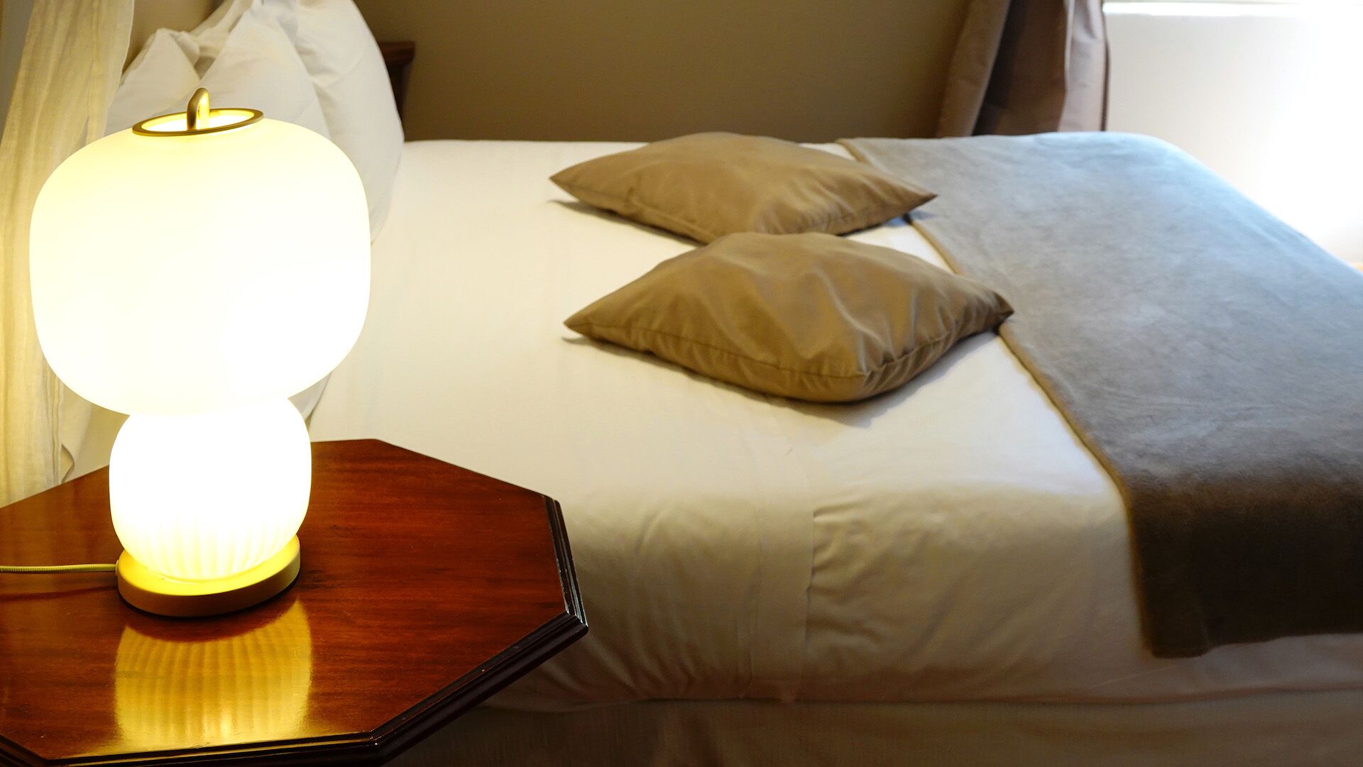 A neatly made bed with white sheets and two beige pillows, a wooden bedside table with a lit lamp, and a window with curtains in the background.
