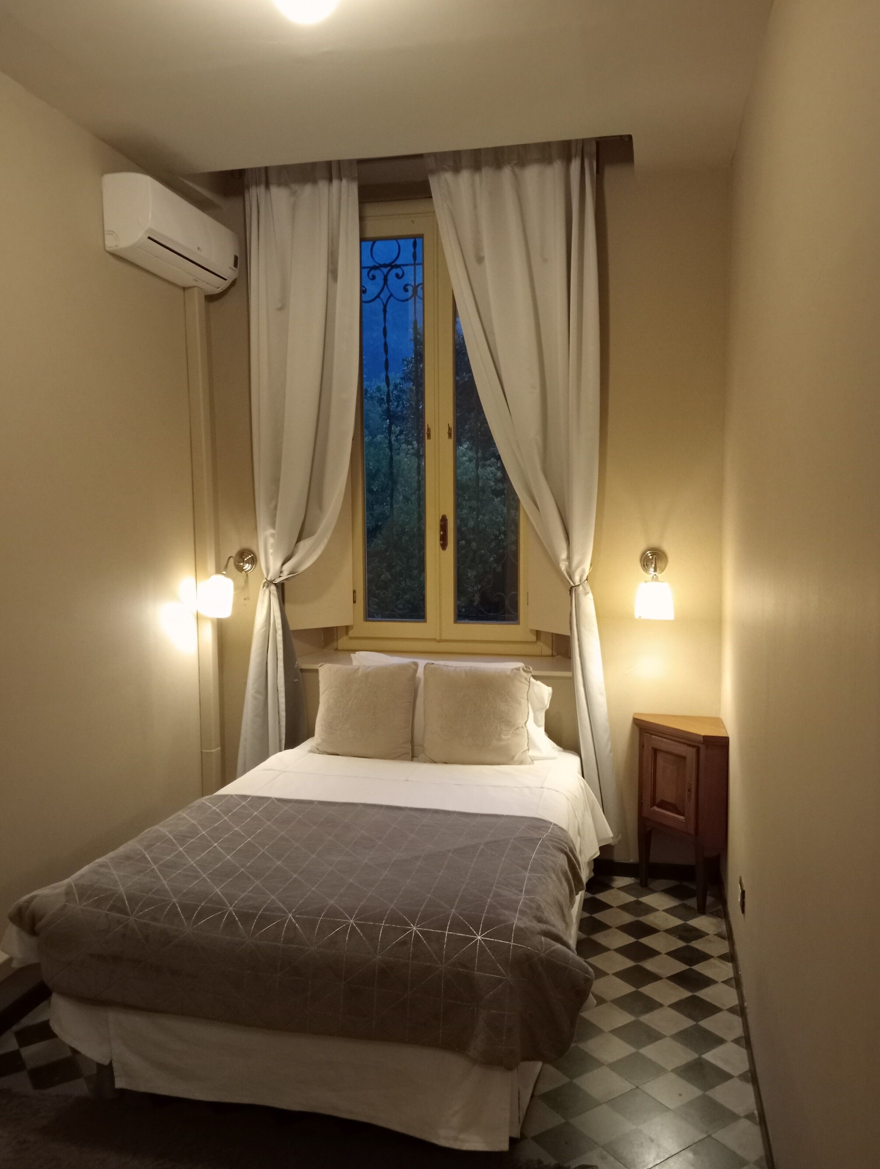 Una camera da letto con un letto, tende, una finestra, un condizionatore d'aria a muro e una lampada da comodino.