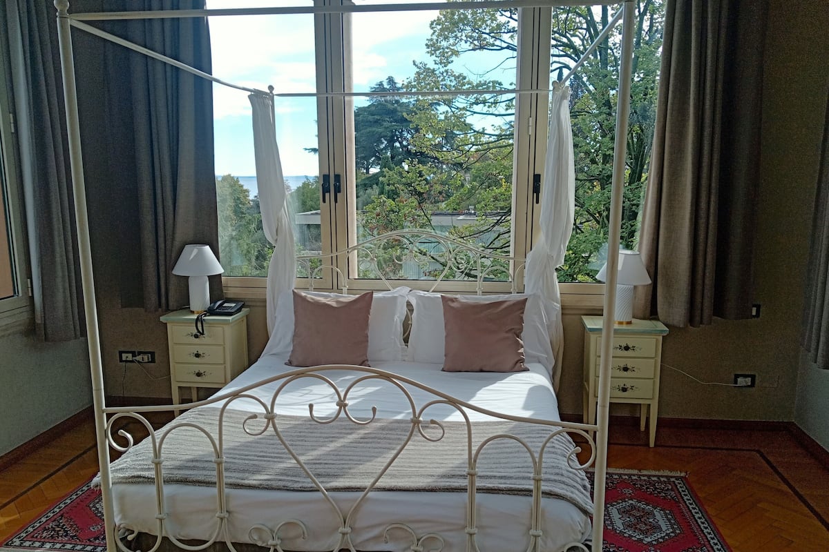 A four-poster canopy bed with a metal frame, two bedside tables with lamps, and a view of trees and sky through large windows.