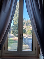 A room with a large window featuring two curtains, a door with a handle, and a view of a landscaped garden with trees and a fence.