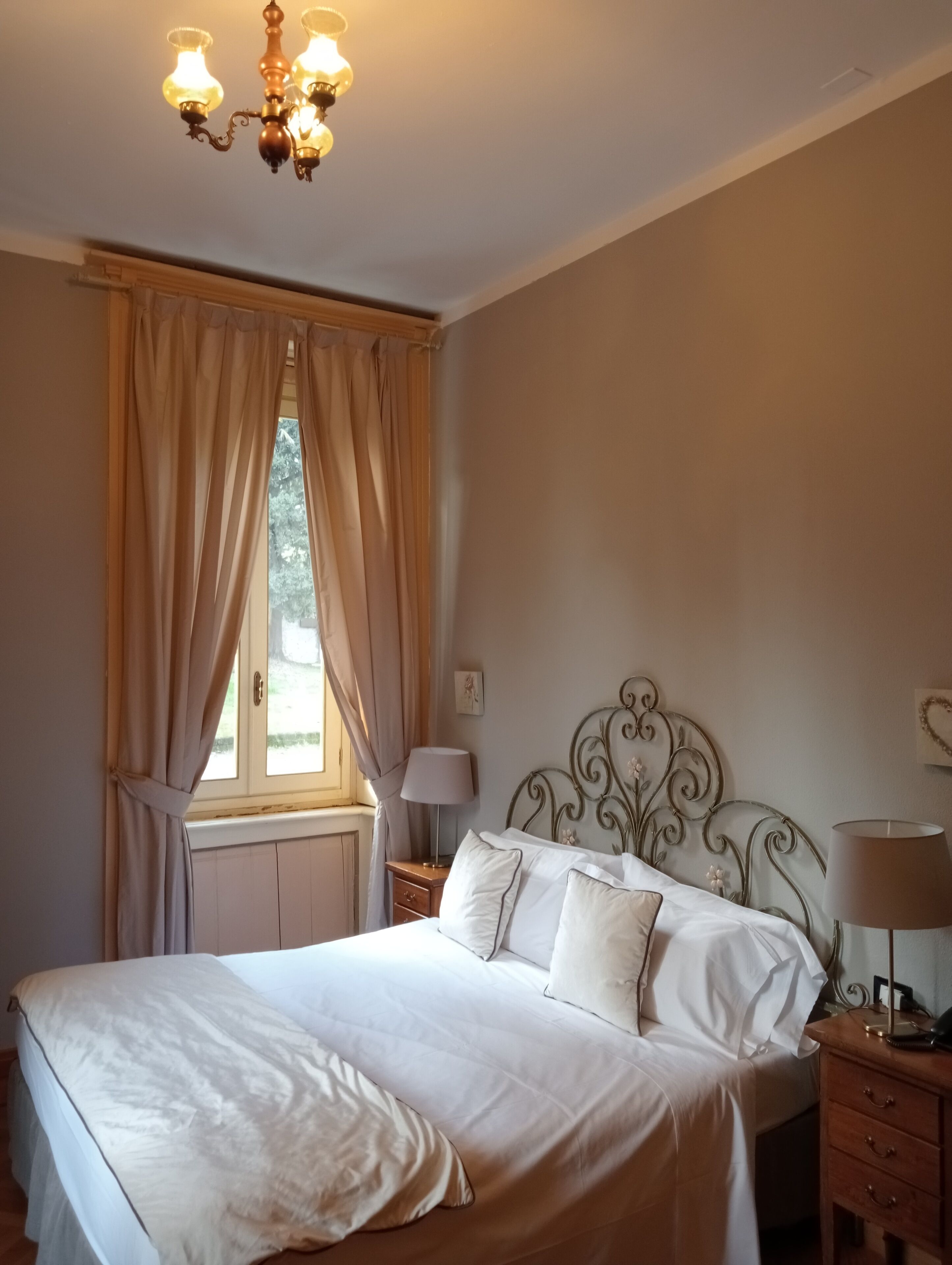Une chambre à coucher avec un grand lit, un lustre, une fenêtre avec des rideaux et une table de chevet avec une lampe.