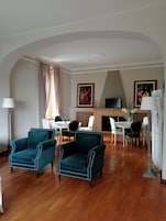 A living room with two teal armchairs, a dining table with chairs, a fireplace, and a television.