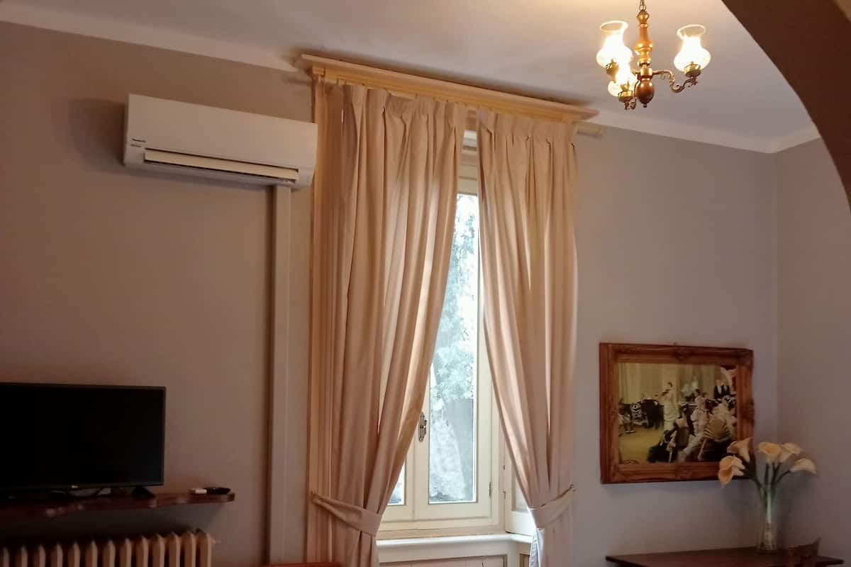 A hotel room with a bed, a chair, a desk with a vase, a painting, an air conditioning unit, and a window with curtains.