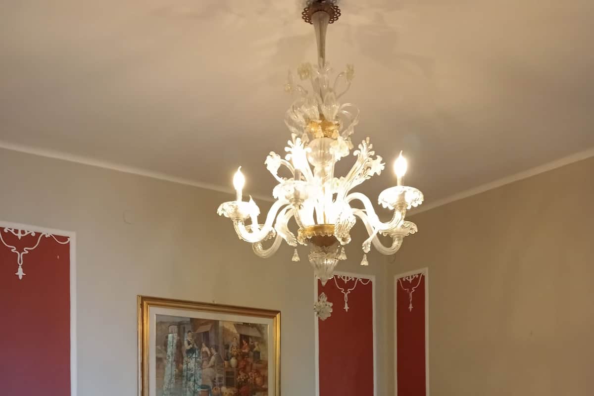 A bedroom with a large bed, a sofa, a chandelier, a fireplace, and a painting on the wall.
