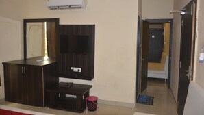A hotel room with a dark wooden wardrobe, a small table, a pink trash bin, and a wall-mounted air conditioner.