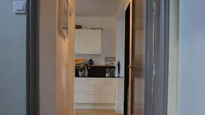 Un couloir étroit, doté d’un plancher en bois, mène à une cuisine avec des meubles blancs et un luminaire suspendu.