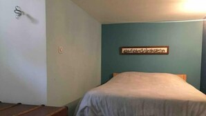 Un lit simple dans une chambre aux murs de couleur turquoise, avec une tête de lit en bois et une couvre-lit de couleur claire. Une valise ancienne est posée sur le sol, à gauche.