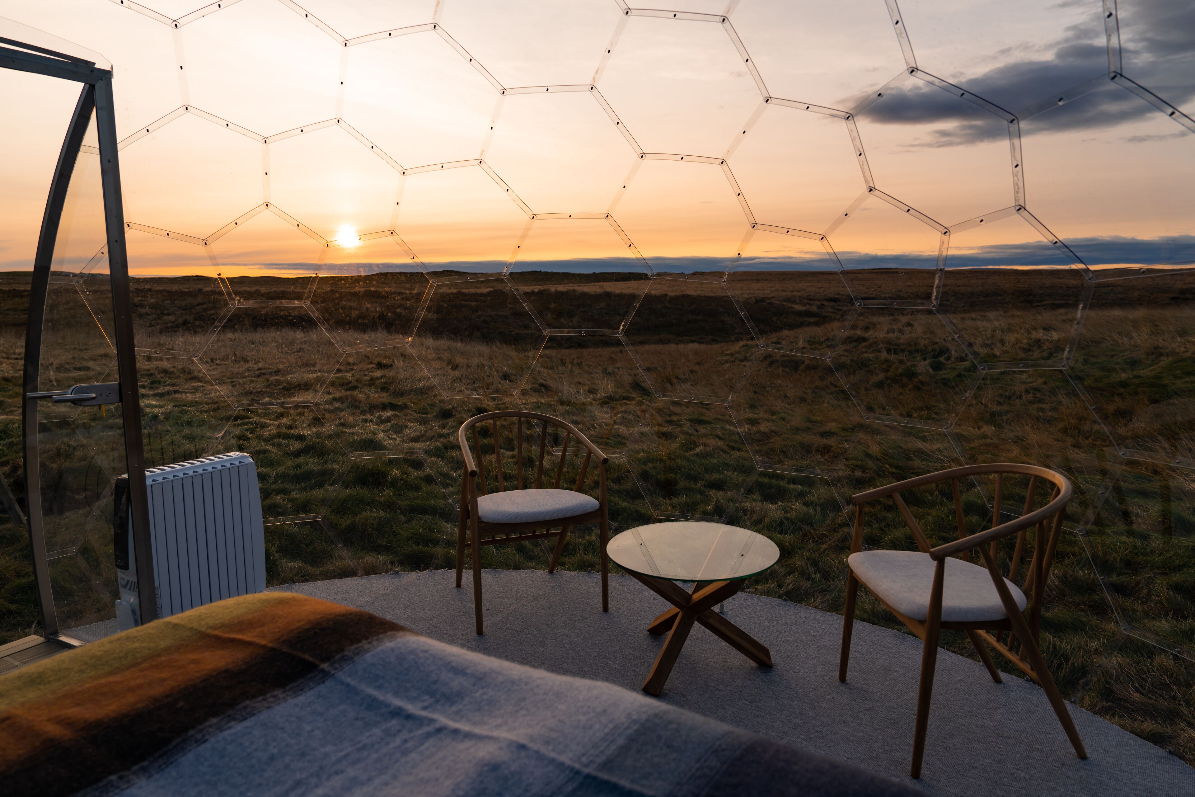 Estructura de vidrio hexagonal con vista al atardecer sobre un campo cubierto de hierba, dos sillas de madera y un radiador.
