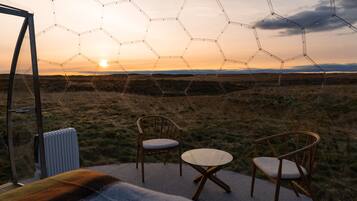 Estructura de vidrio hexagonal con vista al atardecer sobre un campo cubierto de hierba, dos sillas de madera y un radiador.