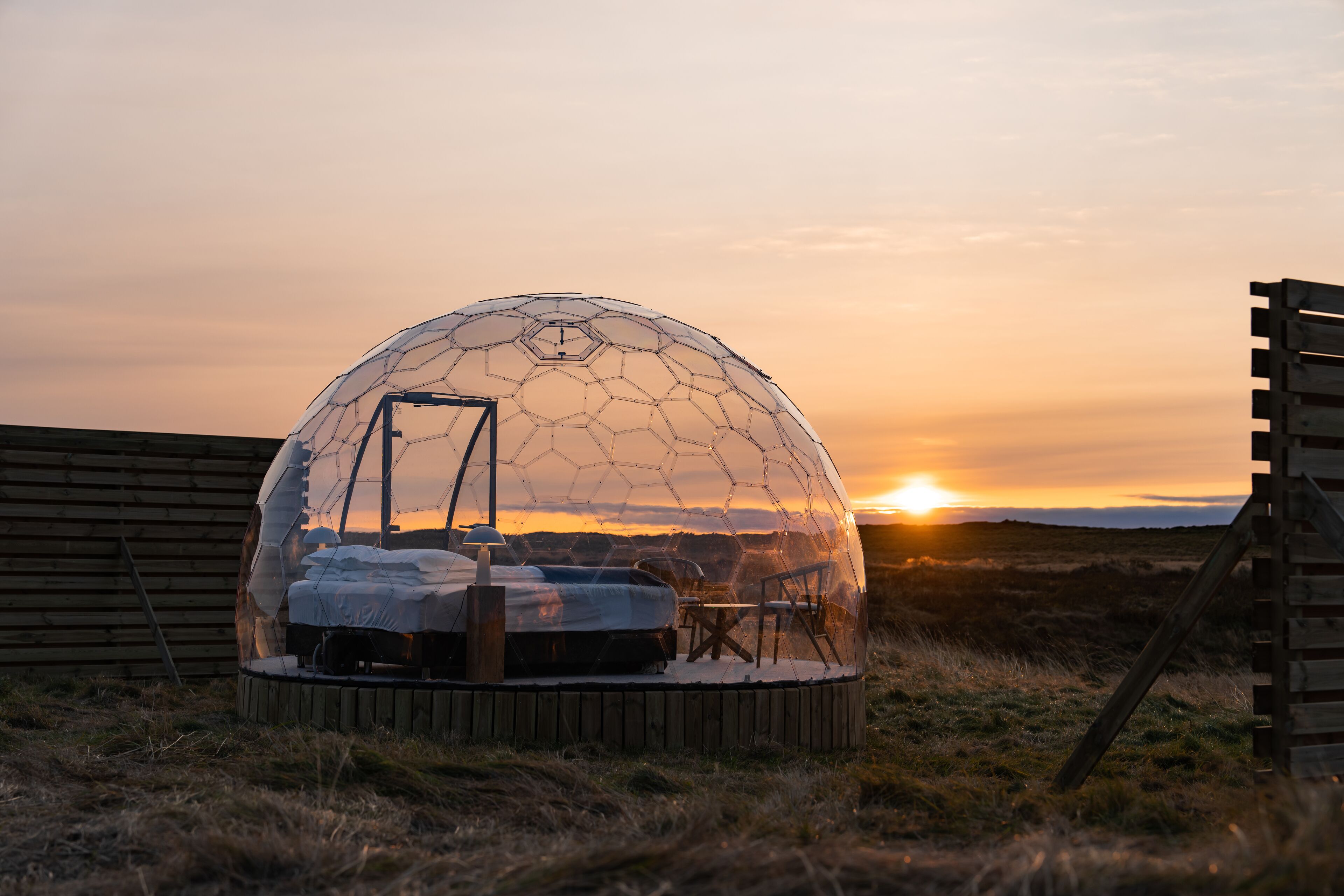 A transparent dome structure with a bed, a table, and chairs inside, set against a sunset background.