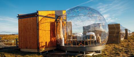 A transparent dome structure adjacent to a wooden building, both situated in an open field.